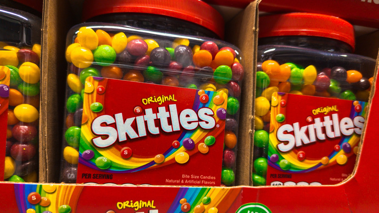 Close up of multiple large containers of rainbow candy with red Skittles label