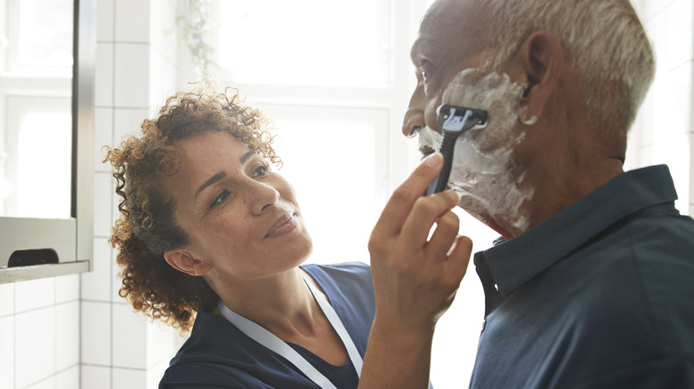 A smiling nurse shaving an elderly man in a bathroom.