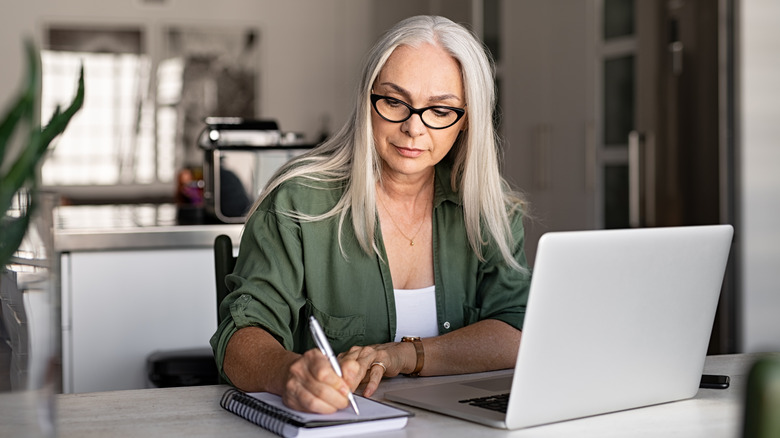 Senior stylish woman taking notes in notebook while using laptop at home.