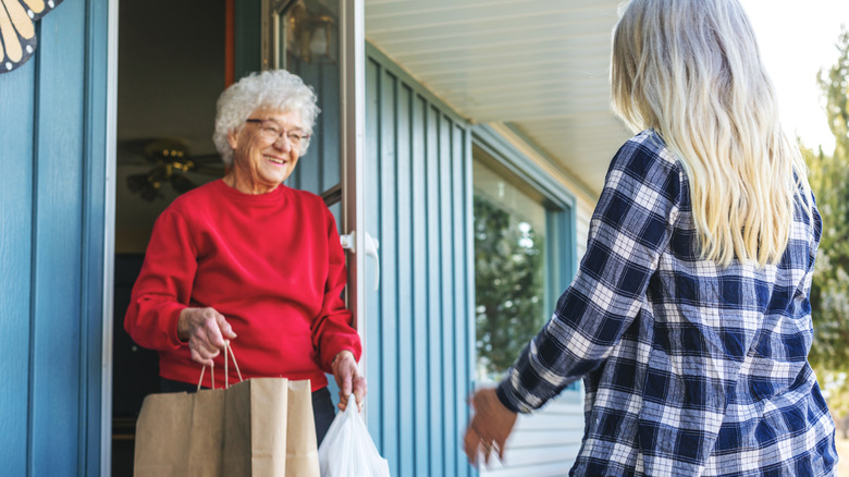 A smiling elderly woman accepting bags of groceries from a younger woman.