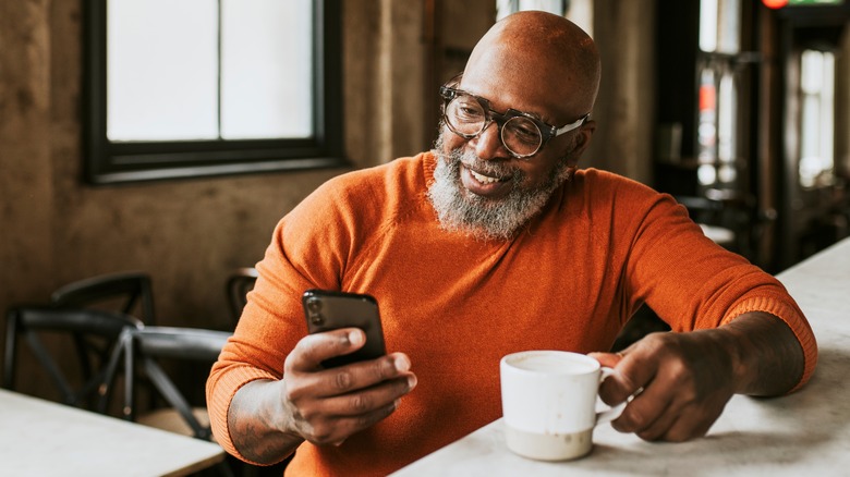 bald bearded man with glasses and holding coffee cup uses phone while seated at coffee bar