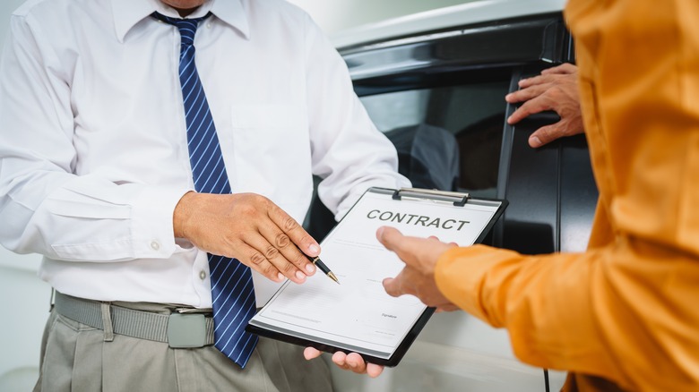 salesman offering contract on clipboard to customer in front of car