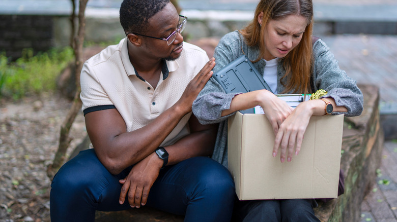 Man and woman sitting in park area, woman is holding box of office items