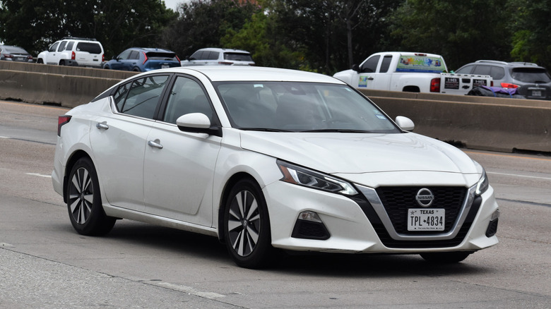 A portrait of a white Nissan Altima traveling down a highway in moderate traffic