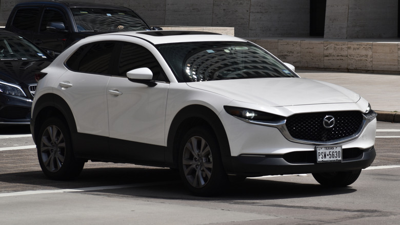 A white Mazda CX-30 Subcompact SUV cruising in a downtown financial district.