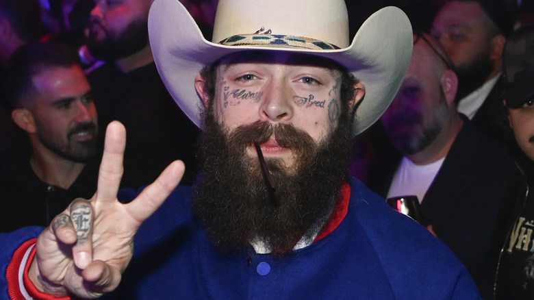 Post Malone wearing white had and smiling while making peace sign