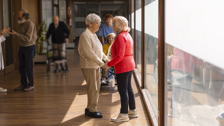 Two elderly women holding hands and talking in a nursing home corridor.