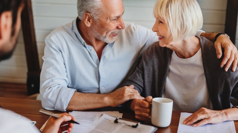 An elderly couple turning and smiling at each other while sitting at a table going over health insurance options with a younger male agent.