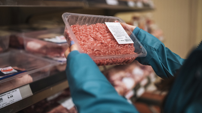 A person holding a package of ground beef with just their arms visible and a shelf of meat in the background