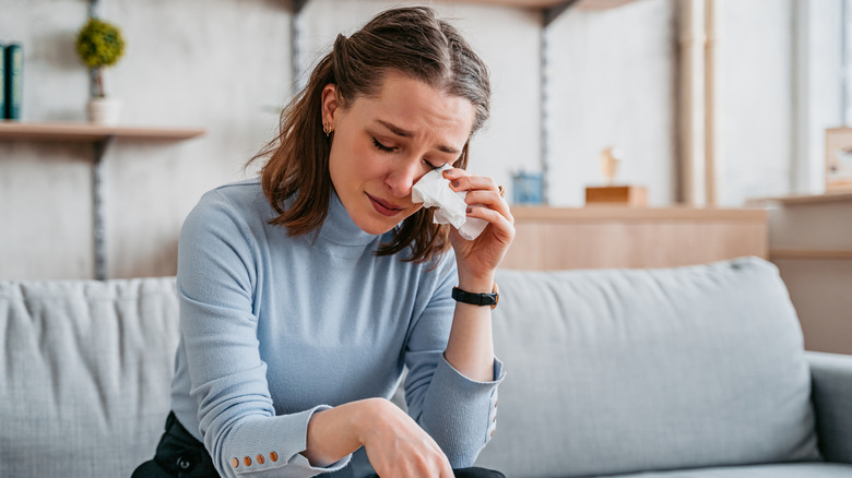 Woman sitting on couch wipes eye while crying