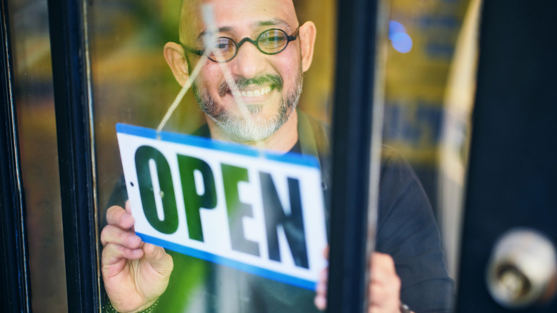 Smiling man wearing eyeglasses behind glass door flipping sign to "open"