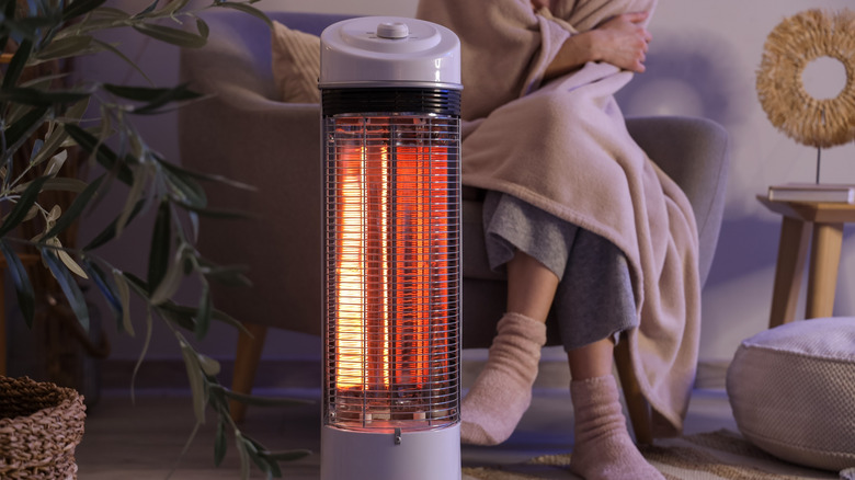 Space heater in foreground of cold woman wrapped in blanket on chair in background