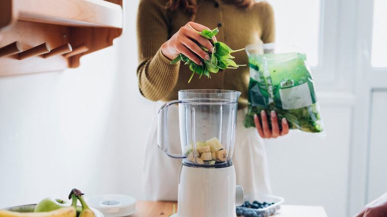 unidentified person in olive shirt putting vegetables into a white blender