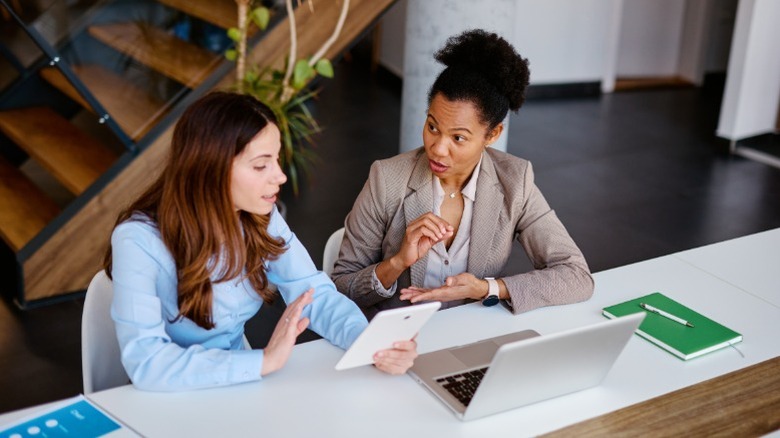 Two women sitting side by side wearing business casual attire and talking