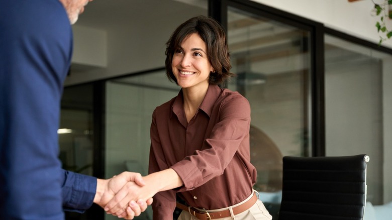 A young woman in a button-down shirt shaking hands with a person in a blue suit inside an office.