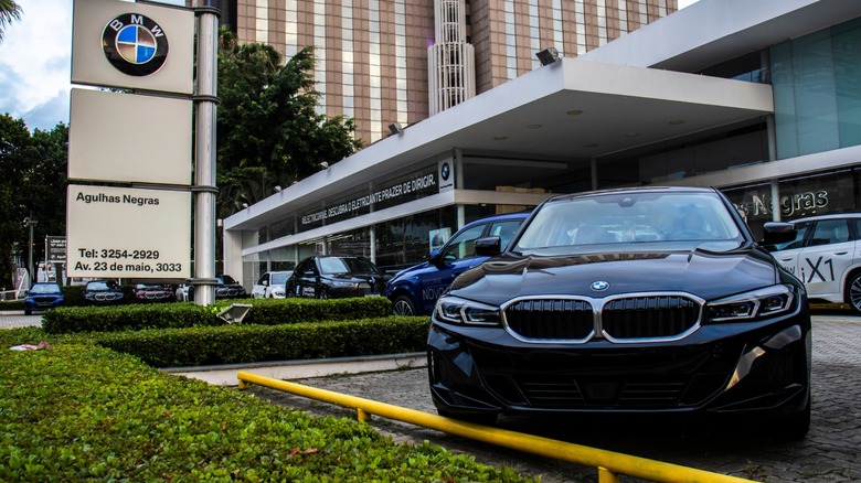Black BMW sedan parked in front of a BMW dealership with showroom building and brand signage visible