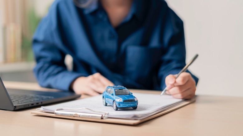 Person signing car paperwork at a desk with a small blue model car on the documents and a laptop nearby