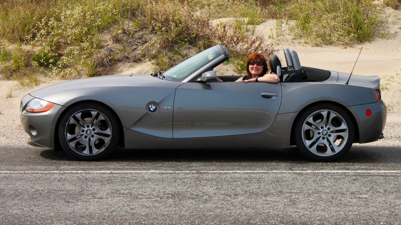 Person smiling in a gray BMW Z4 convertible parked on a roadside near sandy dunes and grass