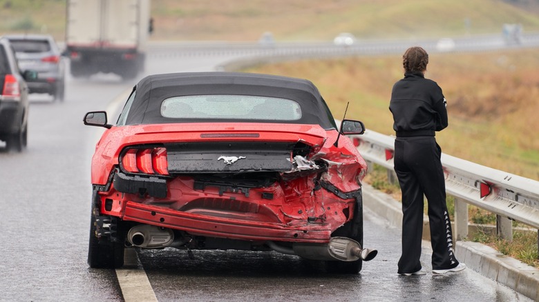 Rear view of a red Ford Mustang convertible with heavy rear-end damage stopped on a wet highway shoulder, with a person standing beside the guardrail