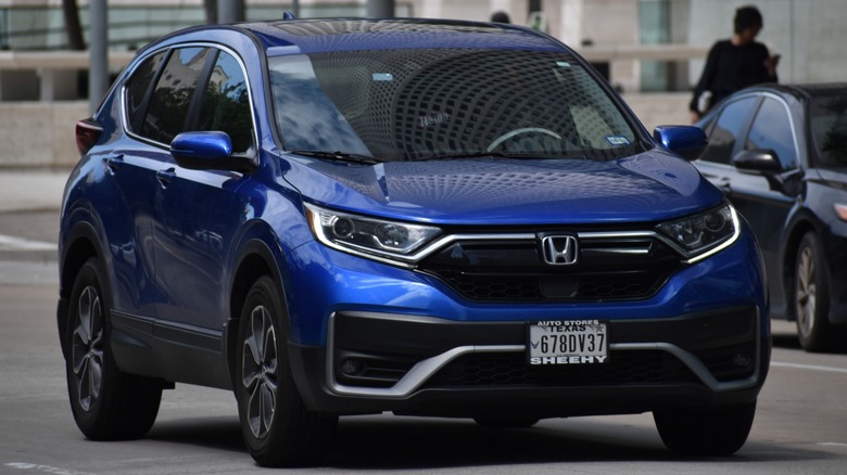 Front three-quarter view of a blue Honda CR-V driving on a city street, with modern buildings reflected on its windshield and hood