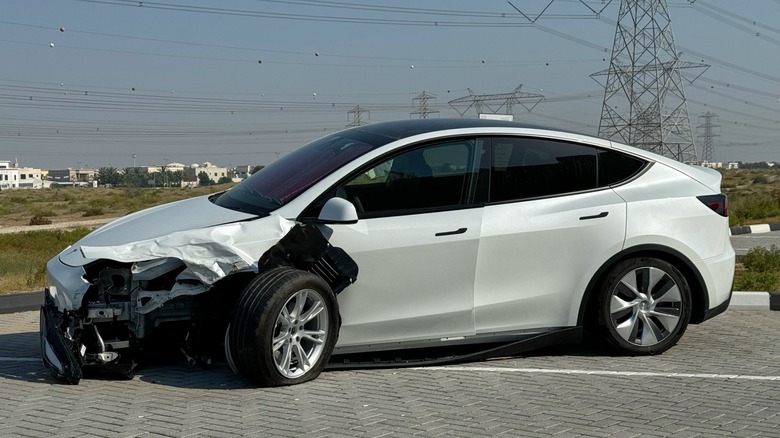White Tesla with significant front-end damage, including a crumpled hood and missing bumper, parked in an outdoor lot