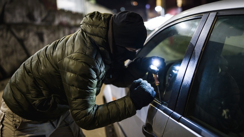 Masked person attempting to break into a parked car at night using a tool
