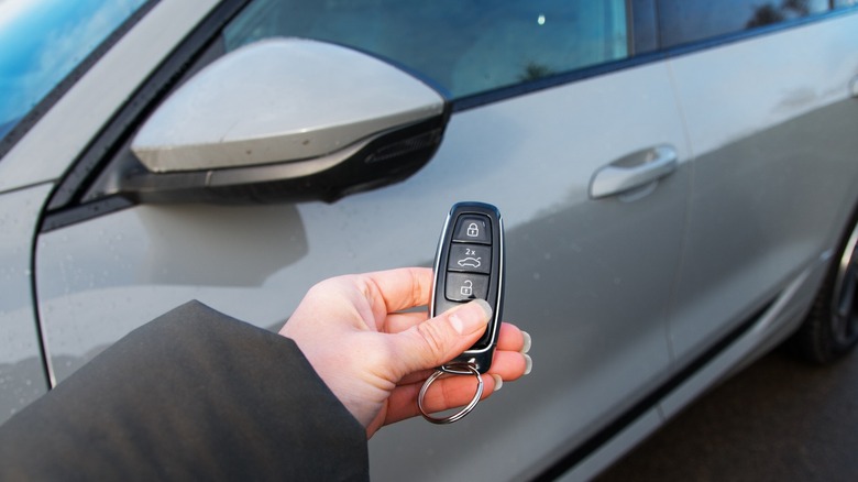 Hand holding a car key fob in front of a parked silver vehicle, with the side mirror and door visible