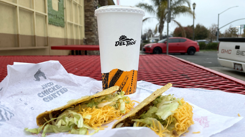 Two tacos and a beverage cup from Del Taco on a table outdoors.