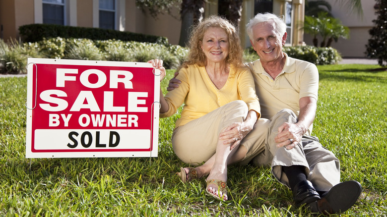 Older couple sit next to a "For Sale By Owner: Sold" sign in front of a home