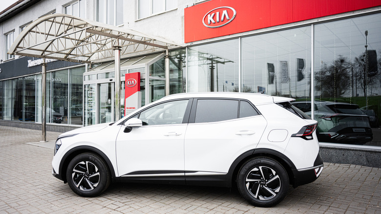 A white 2025 Kia Sportage parked in front of a Kia dealership.