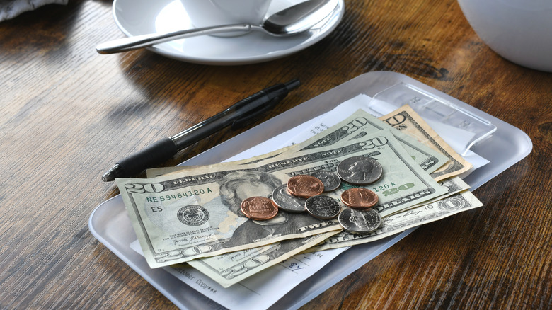 Cash in dollars and coins atop a receipt at a restaurant with a cup, saucer and spoon on the side