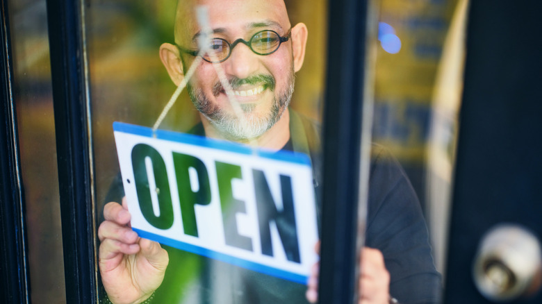 A smiling man wearing glasses flips sign on door to say open
