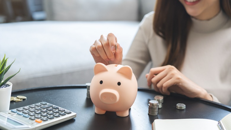A seated woman prepares to put coins into piggy bank next to calculator