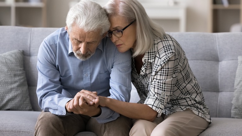 Sad elderly man and women seated on couch holding hands