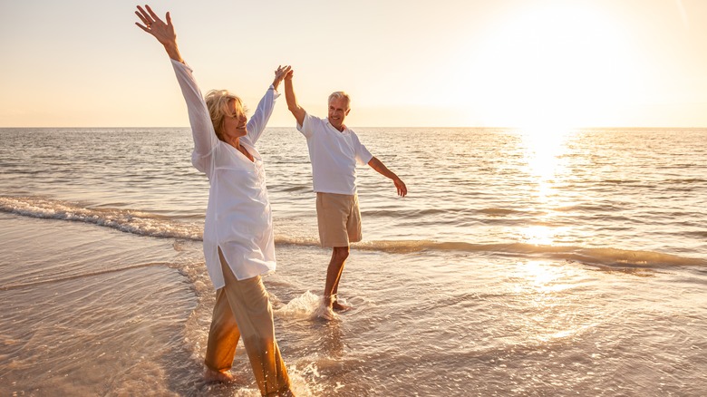 Mature senior couple raise hands in air while waking on sandy beach