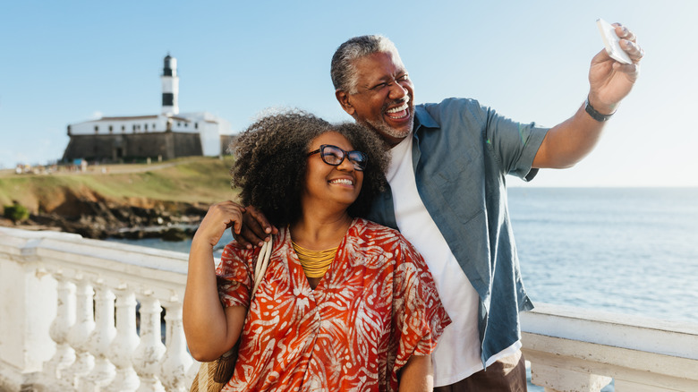 Two seniors smiling taking a selfie in front of a lighthouse near the water.
