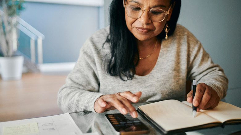 Older woman writing out expenses in a notebook with a calculator