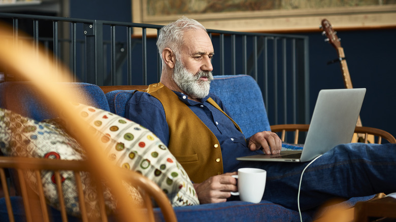 A mature man sitting on a sofa at home with a laptop and a coffee mug