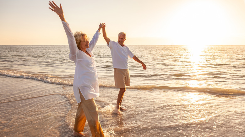 A mature man and woman holding hands while walking barefoot on a beach.