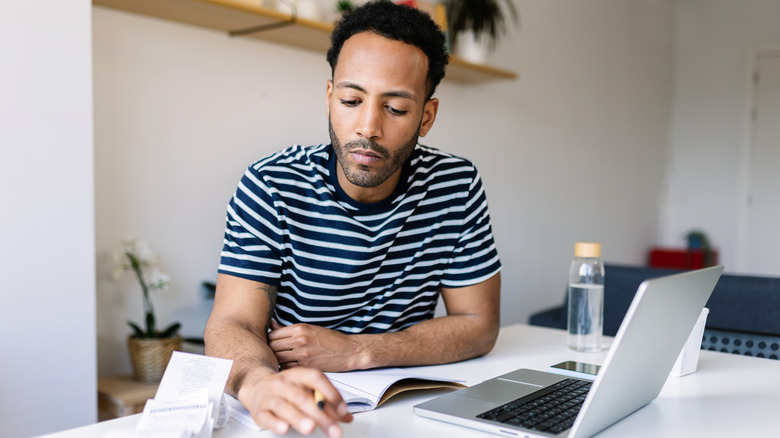 Young adult man with laptop checking bills, taxes, bank account balance and calculating expenses sitting at living room table.