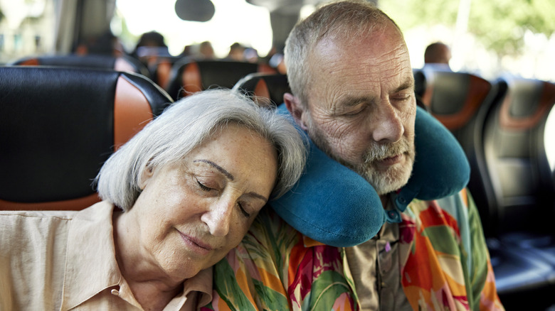 A senior couple asleep on a tour bus with the woman sleeping on the man's shoulder whose wearing a neck pillow.
