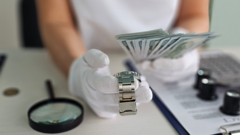 A faceless person with gloved hands examines a watch while holding multiple $100 bills.