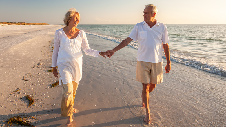 A retired couple holding hands while walking on the beach.