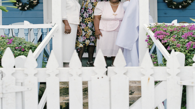 A group of smiling senior women wearing fancy hats on the front porch of a home.