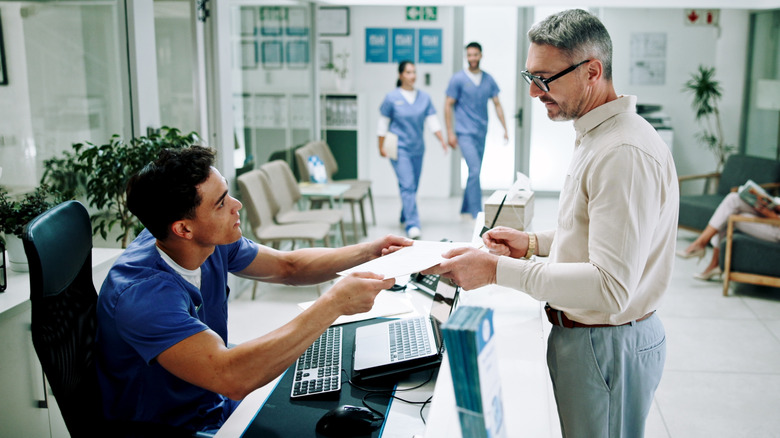 seated ethnically ambiguous male administrative worker hands paperwork to standing white male patient