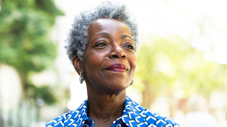 A smiling mature woman standing outside with trees in the background.