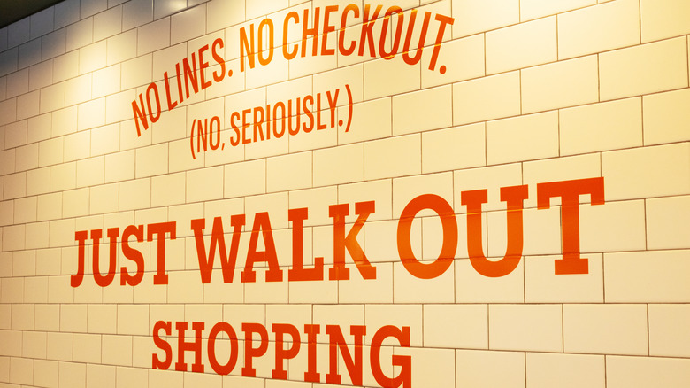 A sign on a white brick wall that says in orange writing "No Lines. No Checkout. (No, Seriously.) Just Walk Out Shopping" in all caps.