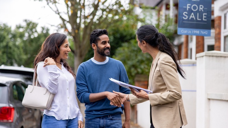 A Millennial couple talking to a realtor outside of a home with a for sale sign on it.