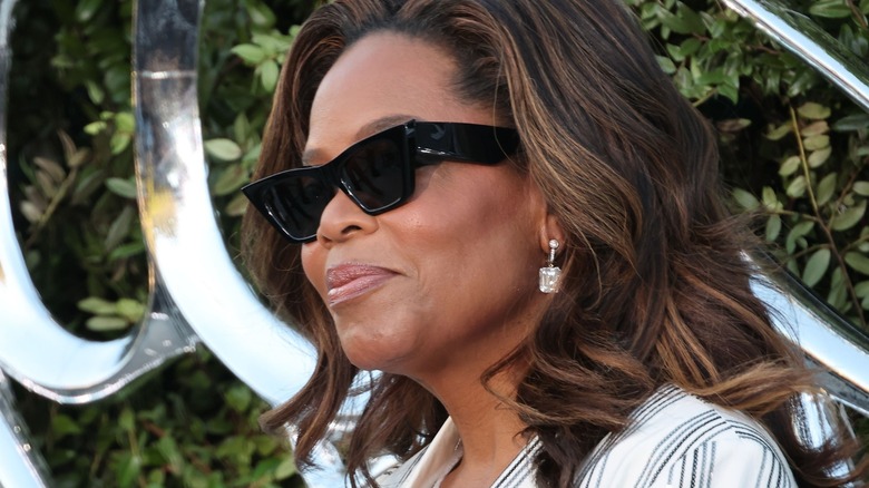 Oprah Winfrey wearing black sunglasses, dangling earrings, and a striped top in front of a leafy backdrop