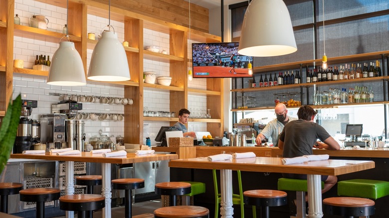 True Food Kitchen restaurant bar with wooden tables, green stools, large pendant lights, and staff serving a seated customerr